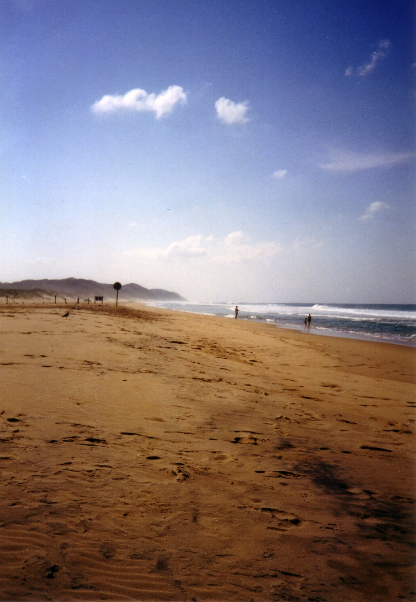 A wide sandy beach at St. Lucia with the Indian Ocean to the right, distant dunes and shoreline in haze, and a few small figures near the water
